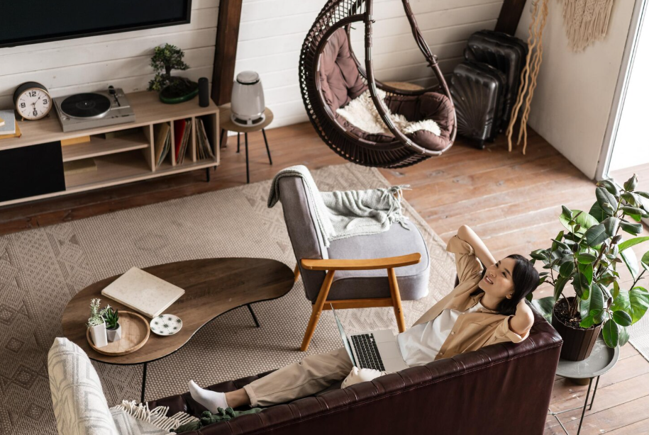 Woman relaxing with a laptop in a cozy clutter living room featuring plants, a hanging chair, vinyl records, and layered textures.