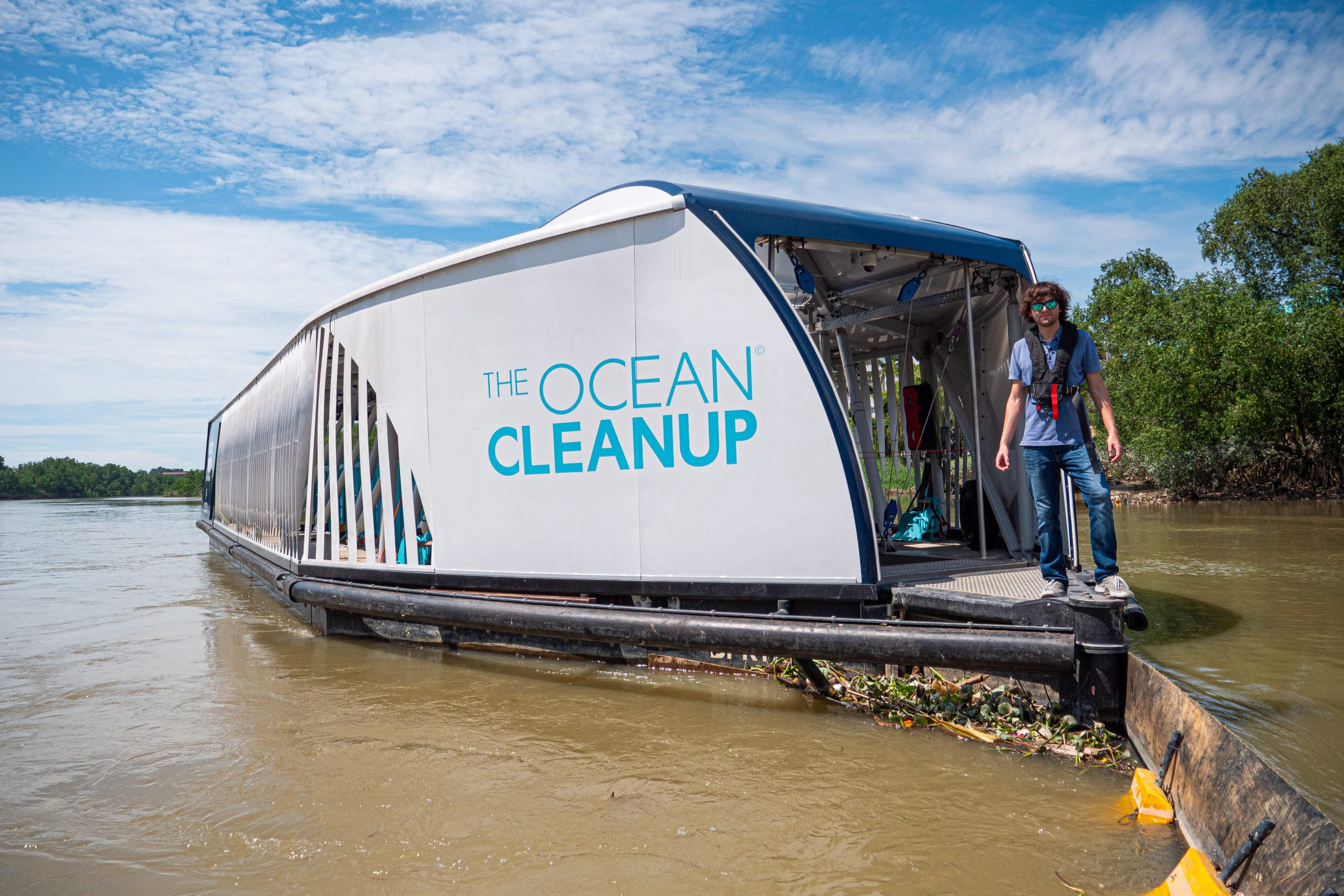 Solar-powered Ocean Cleanup Interceptor vessel collecting river plastic, with a crew member on deck.