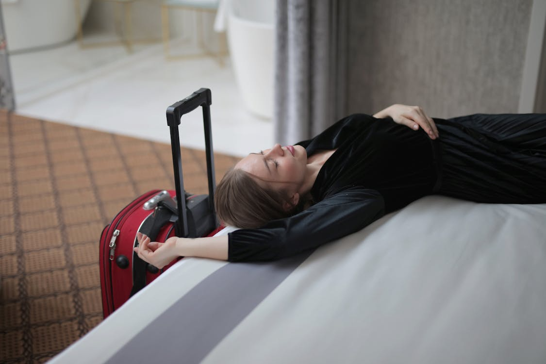 Jet lagged traveler resting on a hotel bed beside a red carry-on suitcase, illustrating travel stress.